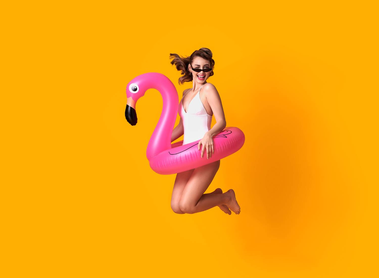 Woman with sunglasses jumping joyfully with a pink flamingo float on a yellow background.
