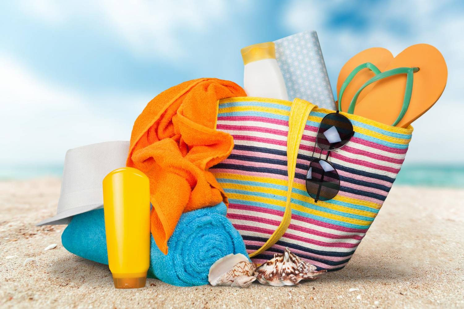 Beach bag with towels, sunscreen, and flip-flops on the sand.