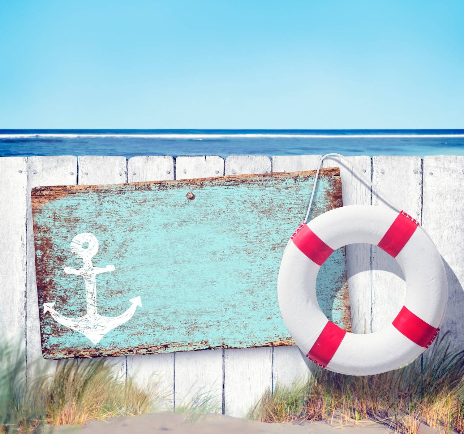 A lifebuoy hangs on a wooden fence with a painted anchor sign near the beach.